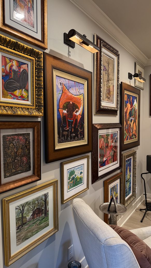 Gallery wall of framed art lit by picture lights above a music room seating area