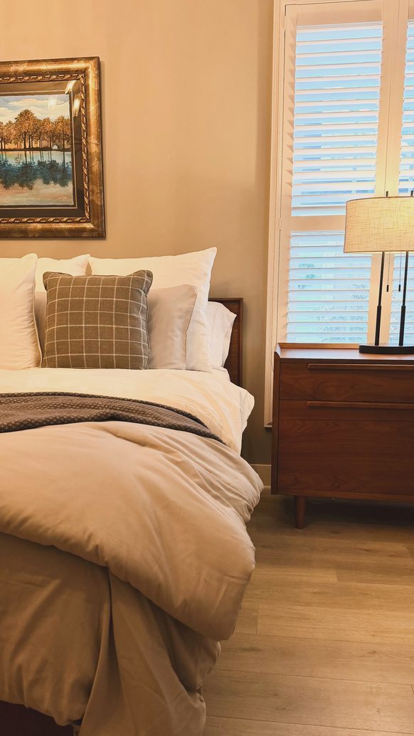 Bedroom with tan and cream bedding, a wood nightstand and framed landscape